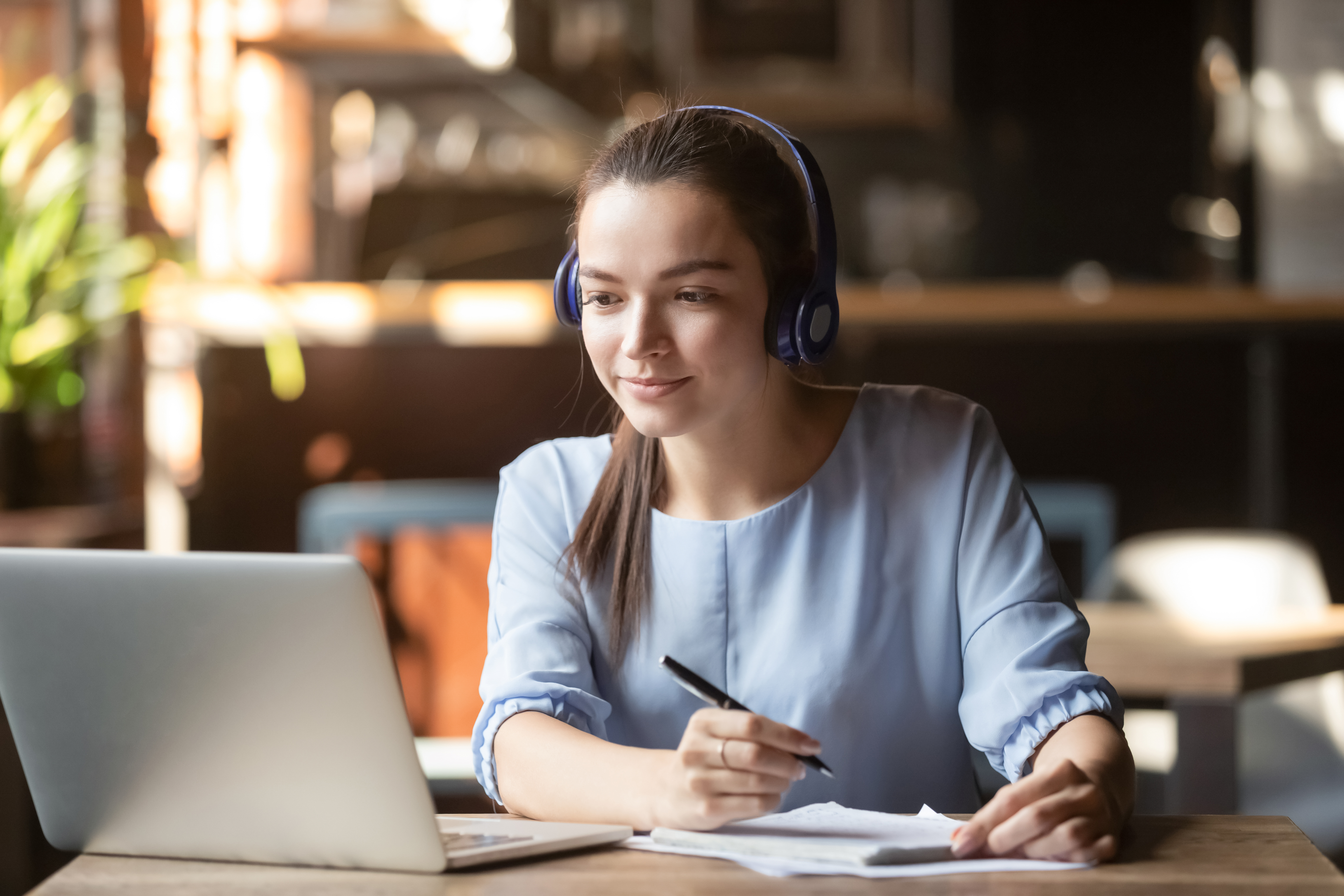 Focused-woman-wearing-headphones-using-laptop,-writing-notes-1152767923_6179x4119